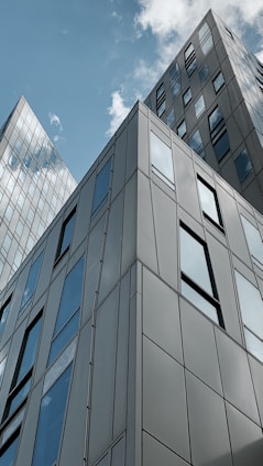 Tall modern buildings with reflective glass windows reach up to a partly cloudy sky. The architecture features clean lines and angular designs, showcasing urban development and contemporary style.