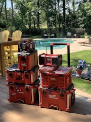 A stack of red Milwaukee Packout toolboxes is neatly arranged on a wooden deck. In the background, there is a clear swimming pool surrounded by patio furniture and trees providing a lush, green environment.
