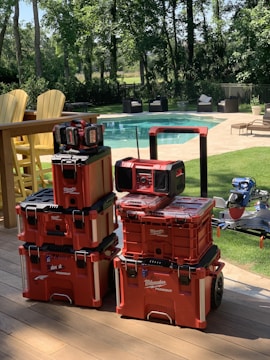 A stack of red Milwaukee Packout toolboxes is neatly arranged on a wooden deck. In the background, there is a clear swimming pool surrounded by patio furniture and trees providing a lush, green environment.