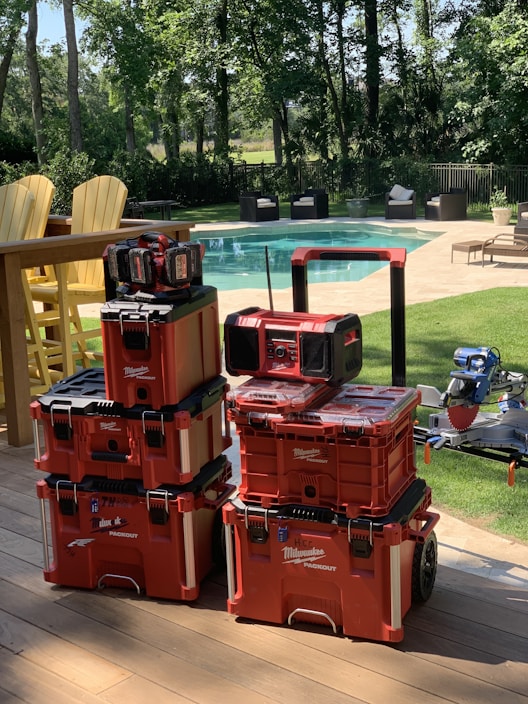 A stack of red Milwaukee Packout toolboxes is neatly arranged on a wooden deck. In the background, there is a clear swimming pool surrounded by patio furniture and trees providing a lush, green environment.