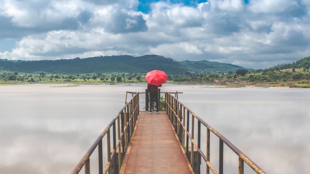 person holding red umbrella walking on wooden bridge