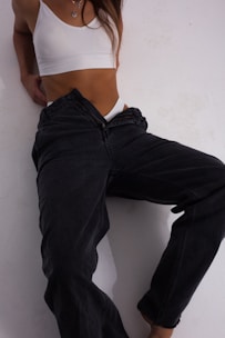 A person is wearing a white crop top and dark jeans, leaning against a white wall. The focus is on the clothing style, with the person's head not visible.