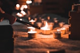 A hand using a lighter to ignite small tealight candles arranged on a textured surface, creating a warm and inviting atmosphere with multiple candles already lit and casting a soft glow.