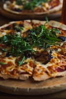 A freshly baked pizza topped with melted cheese, vibrant green arugula, dark olives, and slices of mushrooms, served on a wooden board. The crust appears crispy and golden brown.