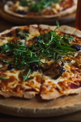 A freshly baked pizza topped with melted cheese, vibrant green arugula, dark olives, and slices of mushrooms, served on a wooden board. The crust appears crispy and golden brown.