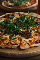 A freshly baked pizza topped with melted cheese, vibrant green arugula, dark olives, and slices of mushrooms, served on a wooden board. The crust appears crispy and golden brown.