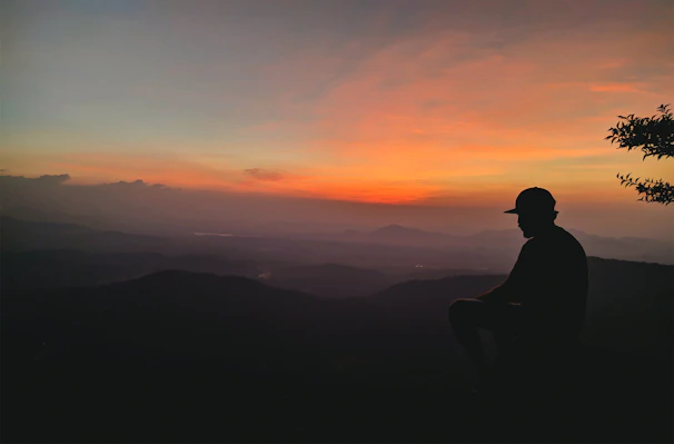 A sunset silhouette of a person wearing a floppy hat, standing on a hilltop.