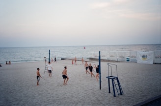 A group of people playing beach volleyball near the water. The scene includes a volleyball net and sand. A calm sea is visible in the background with a few people sitting by the shore, and the sky is overcast with a soft evening light. Advertising banners are placed along the beach fence.
