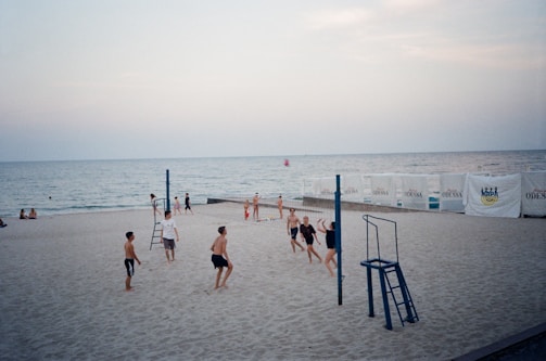A group of people playing beach volleyball near the water. The scene includes a volleyball net and sand. A calm sea is visible in the background with a few people sitting by the shore, and the sky is overcast with a soft evening light. Advertising banners are placed along the beach fence.