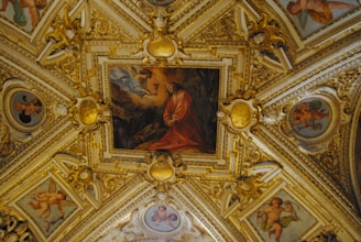 A richly decorated ceiling featuring intricate gold leaf ornamentation and several paintings. The centerpiece is a large painting depicting a biblical or historical scene with figures in robes. Surrounding the main painting are cherubic figures in smaller framed sections, set against a highly detailed background with ornate patterns.