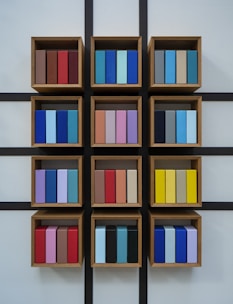 A wall-mounted display of wooden box shelves each containing neatly arranged, colorful rectangular objects. The objects are organized in three rows and four columns with a grid-like black frame. Each shelf holds a variety of colors including shades of blue, red, purple, yellow, and brown.