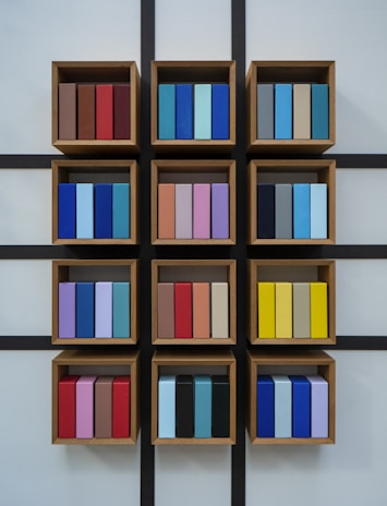 A wall-mounted display of wooden box shelves each containing neatly arranged, colorful rectangular objects. The objects are organized in three rows and four columns with a grid-like black frame. Each shelf holds a variety of colors including shades of blue, red, purple, yellow, and brown.