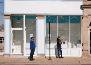 A storefront with white and pastel blue trim, featuring cloud designs printed on the windows. Two individuals stand on the sidewalk in front of the store, both wearing casual clothing and hats. One person wears a red mask and blue jacket, while the other is dressed in a black jacket, holding a white bag, and appears to be using a phone.