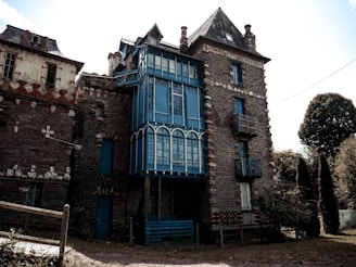 A rustic and slightly aged building with stone and brick construction features a prominent blue accent in its architectural design. The structure has multiple floors, with large windows on the central section, and a distinct triangular roof. The surrounding area includes some trees and foliage, indicating a somewhat secluded location.