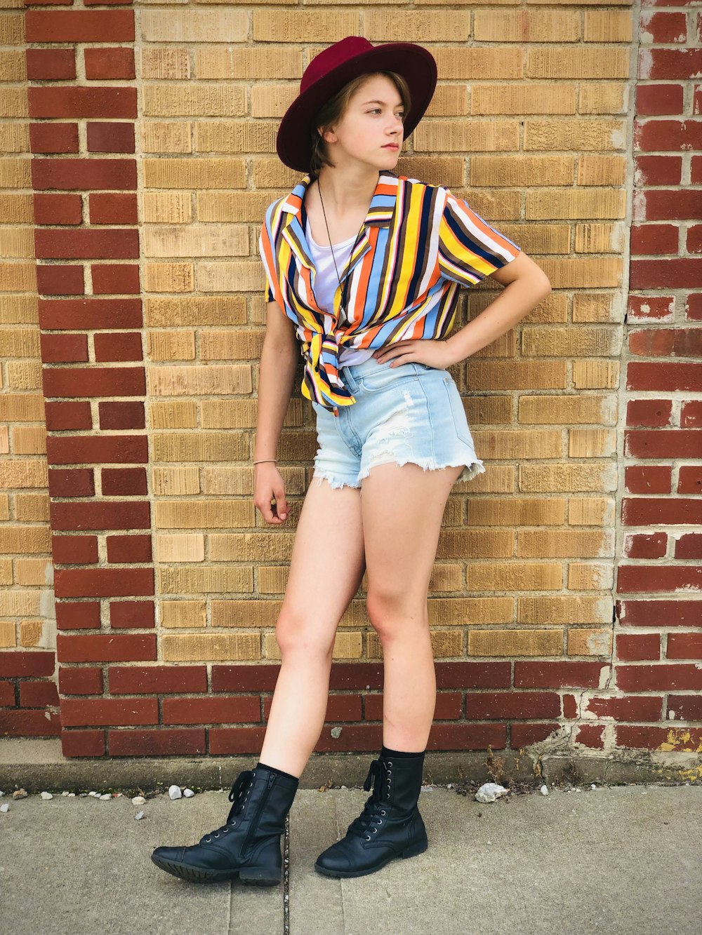 girl in white blue and red stripe shirt and blue denim shorts leaning on brown brick