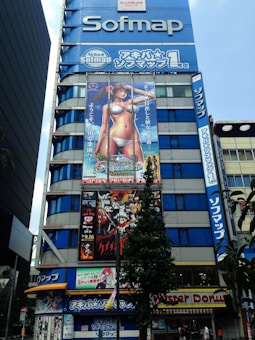 A tall building features multiple large advertisements including anime characters and promotional banners in Japanese. The top of the building displays the Sofmap logo prominently. Below, there are colorful, illustrated banners featuring a woman in a bikini and another with various animated characters. There is foliage and a Mister Donut storefront visible at street level.