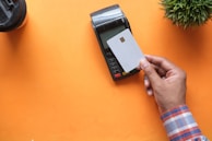 A hand is holding a white credit card near a point-of-sale terminal. The background is an orange surface, and there is a plant and a cup also present in the image.