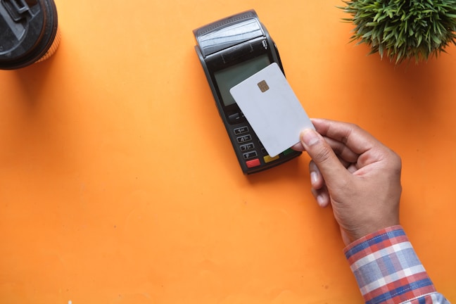 A hand is holding a white credit card near a point-of-sale terminal. The background is an orange surface, and there is a plant and a cup also present in the image.