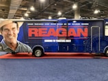 A large promotional vehicle is decorated with an advertisement for a movie titled 'Reagan.' The side features a large image of a smiling man wearing a cowboy hat, next to bold red letters reading 'REAGAN.' Below, in white text, it states 'Coming to Theaters 2022.' The vehicle is on a red carpet, within an indoor setting, with industrial lighting overhead.