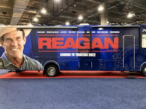 A large promotional vehicle is decorated with an advertisement for a movie titled 'Reagan.' The side features a large image of a smiling man wearing a cowboy hat, next to bold red letters reading 'REAGAN.' Below, in white text, it states 'Coming to Theaters 2022.' The vehicle is on a red carpet, within an indoor setting, with industrial lighting overhead.