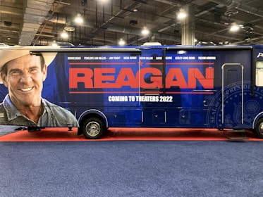 A large promotional vehicle is decorated with an advertisement for a movie titled 'Reagan.' The side features a large image of a smiling man wearing a cowboy hat, next to bold red letters reading 'REAGAN.' Below, in white text, it states 'Coming to Theaters 2022.' The vehicle is on a red carpet, within an indoor setting, with industrial lighting overhead.