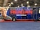 A large promotional vehicle is decorated with an advertisement for a movie titled 'Reagan.' The side features a large image of a smiling man wearing a cowboy hat, next to bold red letters reading 'REAGAN.' Below, in white text, it states 'Coming to Theaters 2022.' The vehicle is on a red carpet, within an indoor setting, with industrial lighting overhead.
