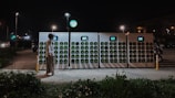 A person wearing a helmet stands before a row of battery charging stations illuminated at night. The charging stations have multiple slots with green indicators, and a small screen is present on each unit. Streetlights are visible in the background, casting light on the scene, along with an area of greenery in the foreground.
