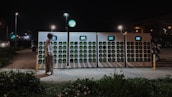 A person wearing a helmet stands before a row of battery charging stations illuminated at night. The charging stations have multiple slots with green indicators, and a small screen is present on each unit. Streetlights are visible in the background, casting light on the scene, along with an area of greenery in the foreground.