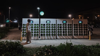 A person wearing a helmet stands before a row of battery charging stations illuminated at night. The charging stations have multiple slots with green indicators, and a small screen is present on each unit. Streetlights are visible in the background, casting light on the scene, along with an area of greenery in the foreground.