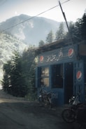 A small, rustic roadside shop with a corrugated metal exterior features several Pepsi logos. The shop is surrounded by dense evergreen trees, set against a backdrop of misty mountains. Two parked motorcycles are visible outside the shop on the dirt road.