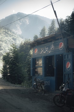 A small, rustic roadside shop with a corrugated metal exterior features several Pepsi logos. The shop is surrounded by dense evergreen trees, set against a backdrop of misty mountains. Two parked motorcycles are visible outside the shop on the dirt road.