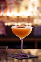 A cocktail glass filled with an orange-hued drink, garnished with a slice of passion fruit on top. The glass sits on a dark napkin on a reflective surface, with a blurred, warmly lit bar background.