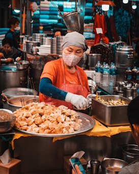 A vendor wearing a face mask and gloves serves food at a busy market stall. The scene features steaming pots, trays of dumplings, and an assortment of kitchen utensils. Colorful fabrics are stacked in the background, creating a vibrant atmosphere.