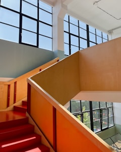 A modern, spacious staircase with bright red steps and light wooden railings is illuminated by large windows, allowing natural light to flood the space. The walls are painted in soft, neutral colors, and outdoor greenery is visible through the glass.