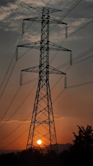 A large metal electricity pylon stands tall against the backdrop of a sunset, with the sun positioned near the base of the structure. The sky shows a mix of dark clouds and orange hues, creating a dramatic silhouette effect. Power lines extend outward from the pylon, partially visible against the sky. Trees and bushes are present in the lower part of the image.