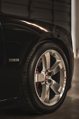 A close-up view of a car's chrome alloy wheel and tire, featuring a logo at the center. The wheel is shiny and well-maintained, with visible tire tread. A badge labeled 'HEMI' is visible on the car's dark paintwork, indicating a high-performance engine.