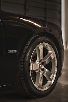A close-up view of a car's chrome alloy wheel and tire, featuring a logo at the center. The wheel is shiny and well-maintained, with visible tire tread. A badge labeled 'HEMI' is visible on the car's dark paintwork, indicating a high-performance engine.
