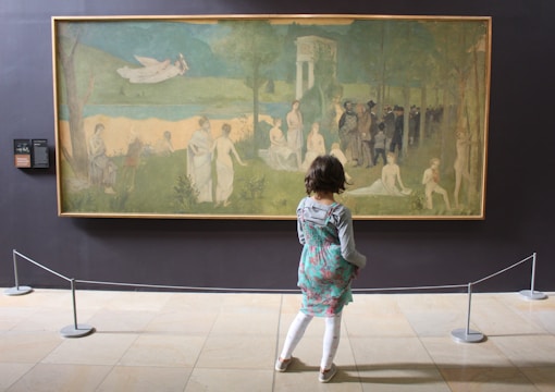 A child is standing in front of a large artwork displayed in a gallery. The painting depicts a serene, pastoral scene with numerous figures in white robes scattered across a grassy landscape. There is an ethereal atmosphere, with a winged figure flying in the sky and a group of people gathered in a wooded area. The scene is framed by trees and classical architecture.