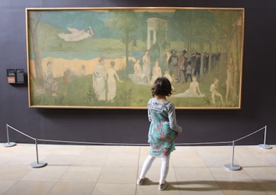 A child is standing in front of a large artwork displayed in a gallery. The painting depicts a serene, pastoral scene with numerous figures in white robes scattered across a grassy landscape. There is an ethereal atmosphere, with a winged figure flying in the sky and a group of people gathered in a wooded area. The scene is framed by trees and classical architecture.