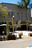 A street view captures an outdoor seating area adjacent to a building marked 'Realty Building.' The exterior includes several metal kegs and wooden planters with small trees lining the sidewalk. A saloon with a prominent sign is partially visible, surrounded by greenery and outdoor furniture.