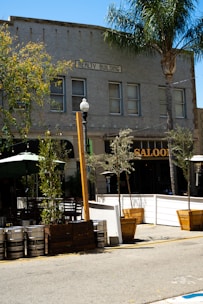 A street view captures an outdoor seating area adjacent to a building marked 'Realty Building.' The exterior includes several metal kegs and wooden planters with small trees lining the sidewalk. A saloon with a prominent sign is partially visible, surrounded by greenery and outdoor furniture.