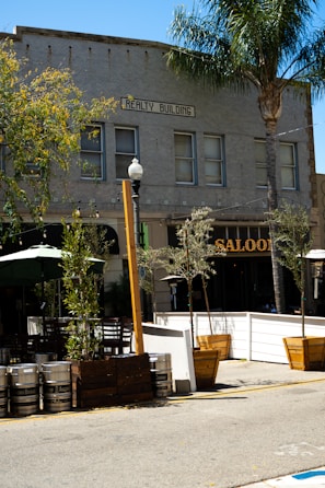 A street view captures an outdoor seating area adjacent to a building marked 'Realty Building.' The exterior includes several metal kegs and wooden planters with small trees lining the sidewalk. A saloon with a prominent sign is partially visible, surrounded by greenery and outdoor furniture.