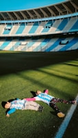 Two children are lying on the grass of a large, empty football stadium. They are wearing matching striped sports jerseys, and the sunlight casts shadows on the field. The stadium seating is painted in alternating blue and white sections.