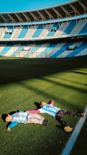 Two children are lying on the grass of a large, empty football stadium. They are wearing matching striped sports jerseys, and the sunlight casts shadows on the field. The stadium seating is painted in alternating blue and white sections.