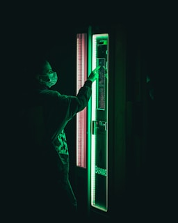 A person wearing a mask is interacting with a vending machine that emits a green glow. The scene is dark with the green light being the focal point.