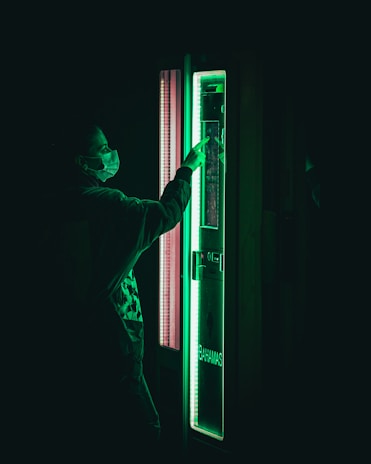 A person wearing a mask is interacting with a vending machine that emits a green glow. The scene is dark with the green light being the focal point.