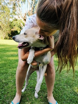 A person with long hair embraces a light-colored dog outdoors on a grassy area surrounded by trees. The dog appears content and relaxed, with its tongue out. The person's posture shows affection as they hold the dog close.