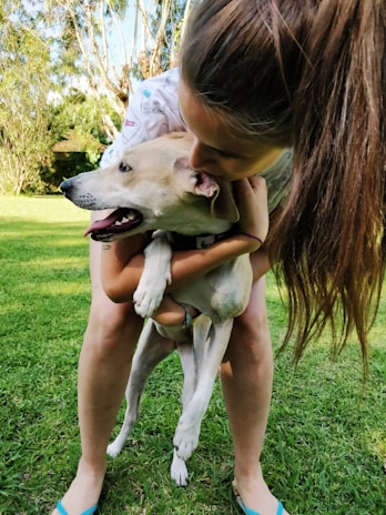 A person with long hair embraces a light-colored dog outdoors on a grassy area surrounded by trees. The dog appears content and relaxed, with its tongue out. The person's posture shows affection as they hold the dog close.