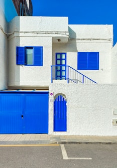 A white stucco building with vibrant blue doors, shutters, and trim creates a striking contrast. The two-story structure features an upper balcony with blue railing and a large blue garage door below. The building is positioned on a paved street with a sidewalk in front.