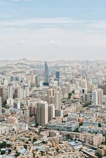 A sprawling cityscape with numerous high-rise buildings and residential complexes. The urban landscape features a mix of modern skyscrapers and older structures, set against a backdrop of distant hills under a clear blue sky.